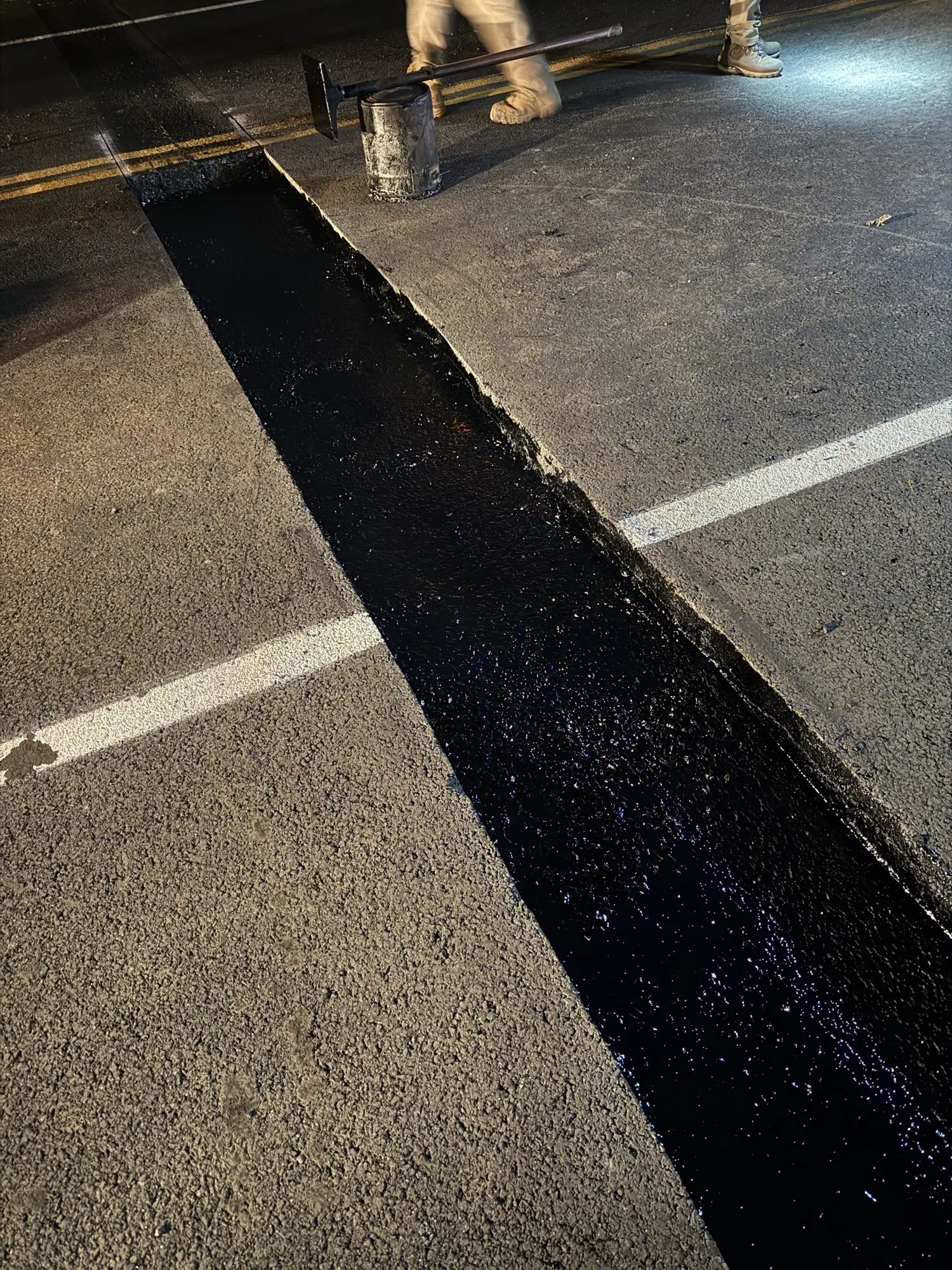 Worker applying hot rubberized crack sealant across a highway lane at night