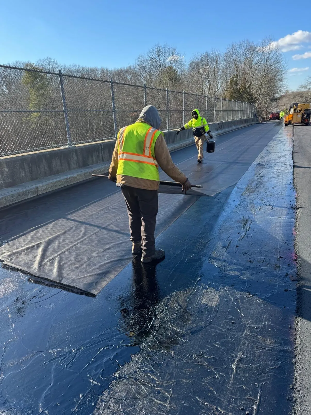 Worker rolling out and squeegeeing sheet waterproofing membrane along a bridge deck between chain link fencing