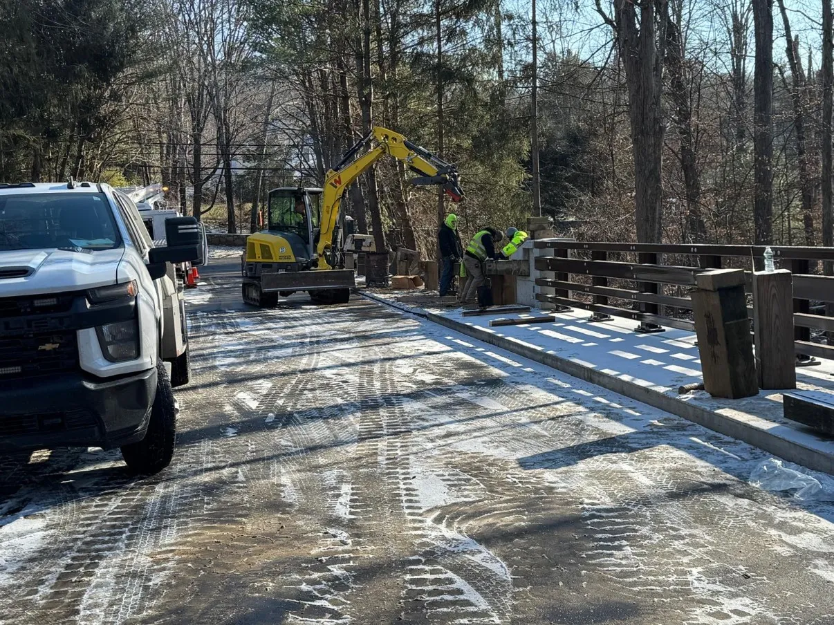 Completed bridge deck after washing and sealing, with new concrete parapet and steel railing posts and an excavator working the far abutment