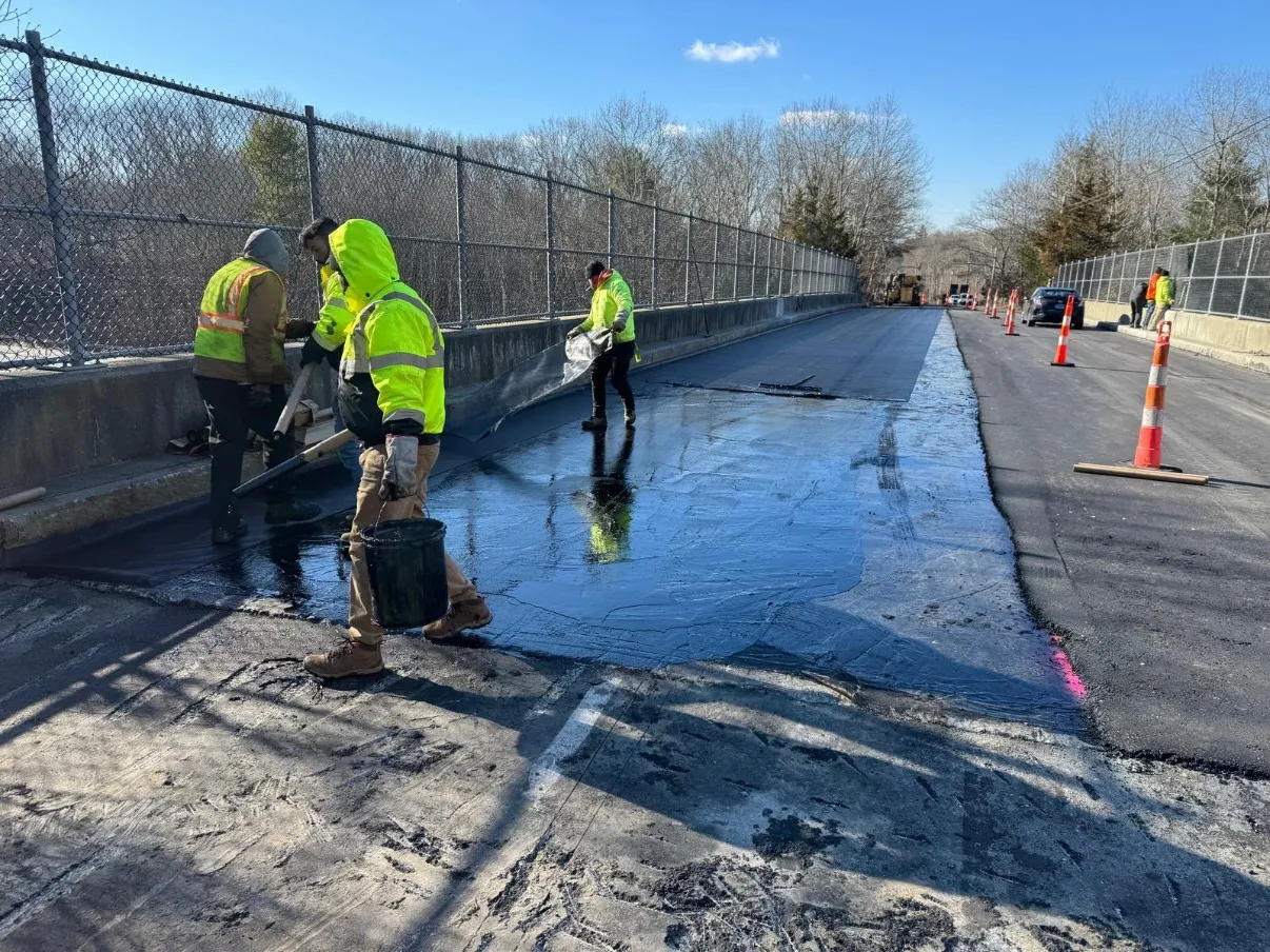 Cristiano & Son crew in high-visibility gear applying black waterproofing membrane across a fenced bridge deck ahead of paving