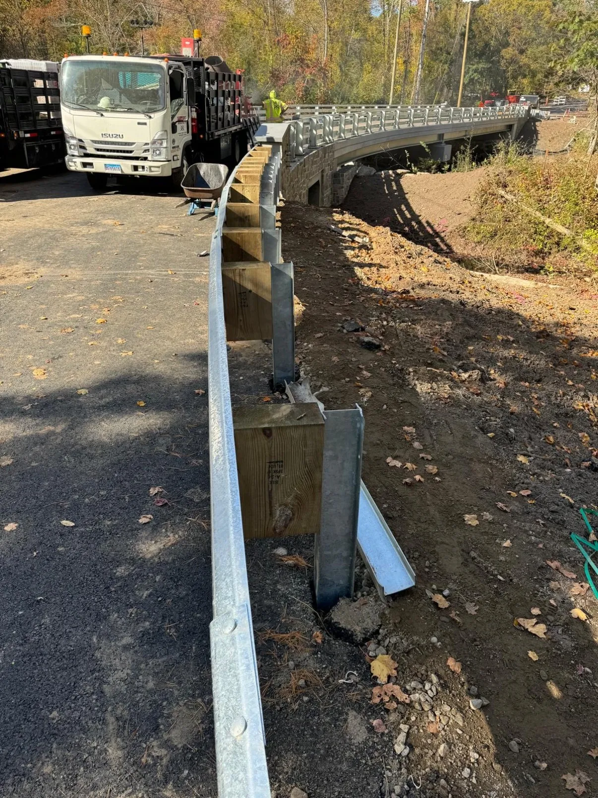 Guardrail run curving down a paved bridge approach into a concrete anchor block, with a flatbed truck of W-beam rail staged alongside