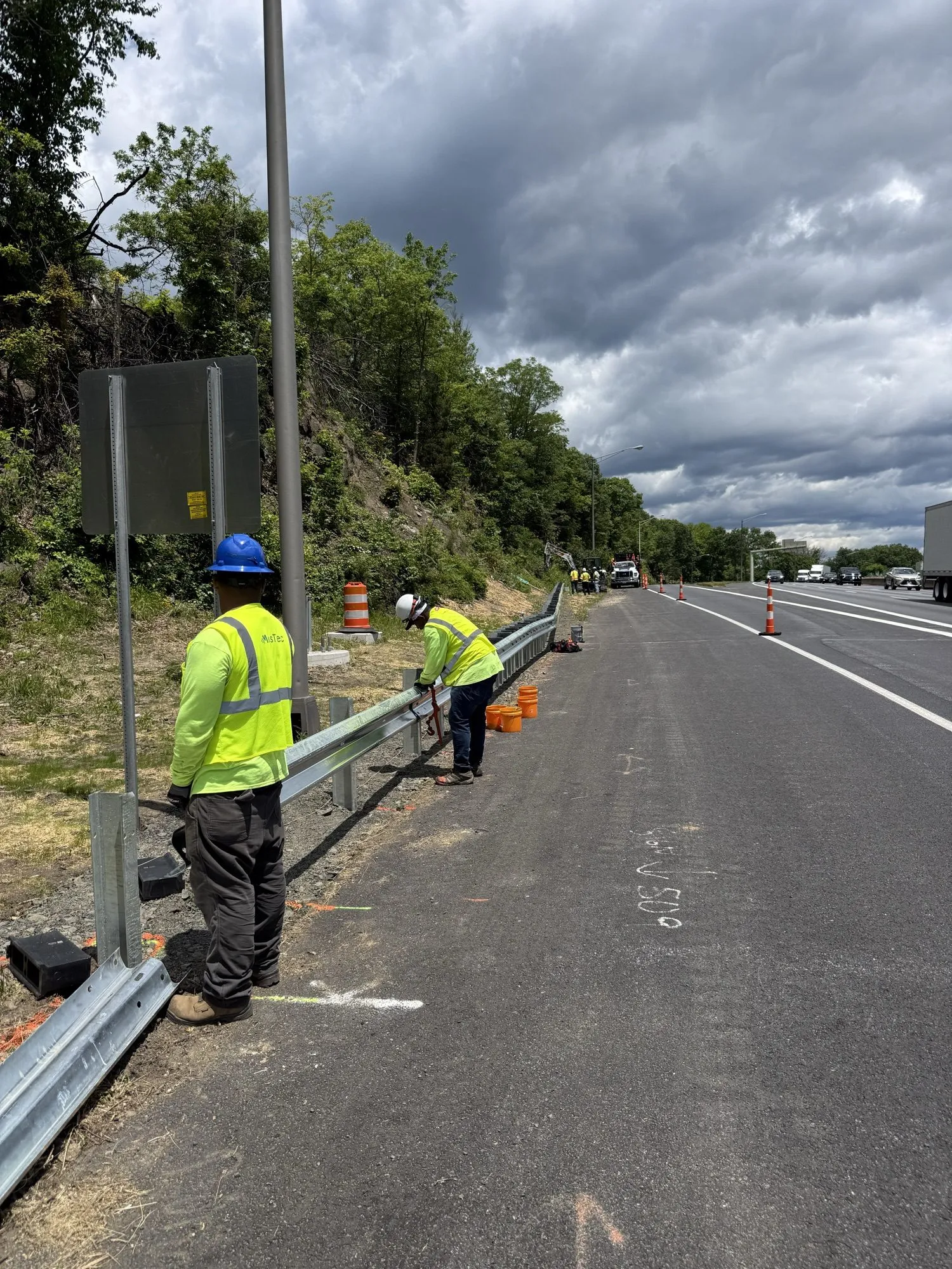 Crew bolting W-beam guardrail on a highway shoulder under storm clouds