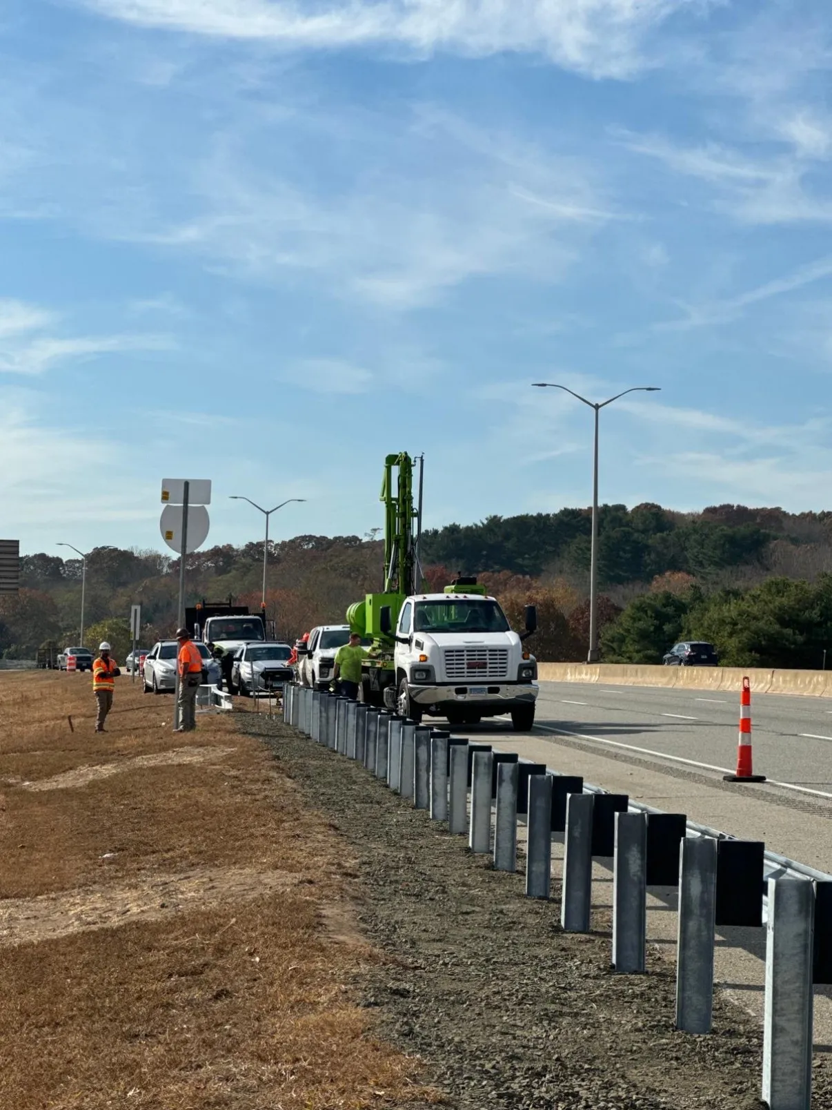 Line of newly driven guardrail posts along a divided highway shoulder with a Cristiano & Son crew, cones, and a truck-mounted post driver