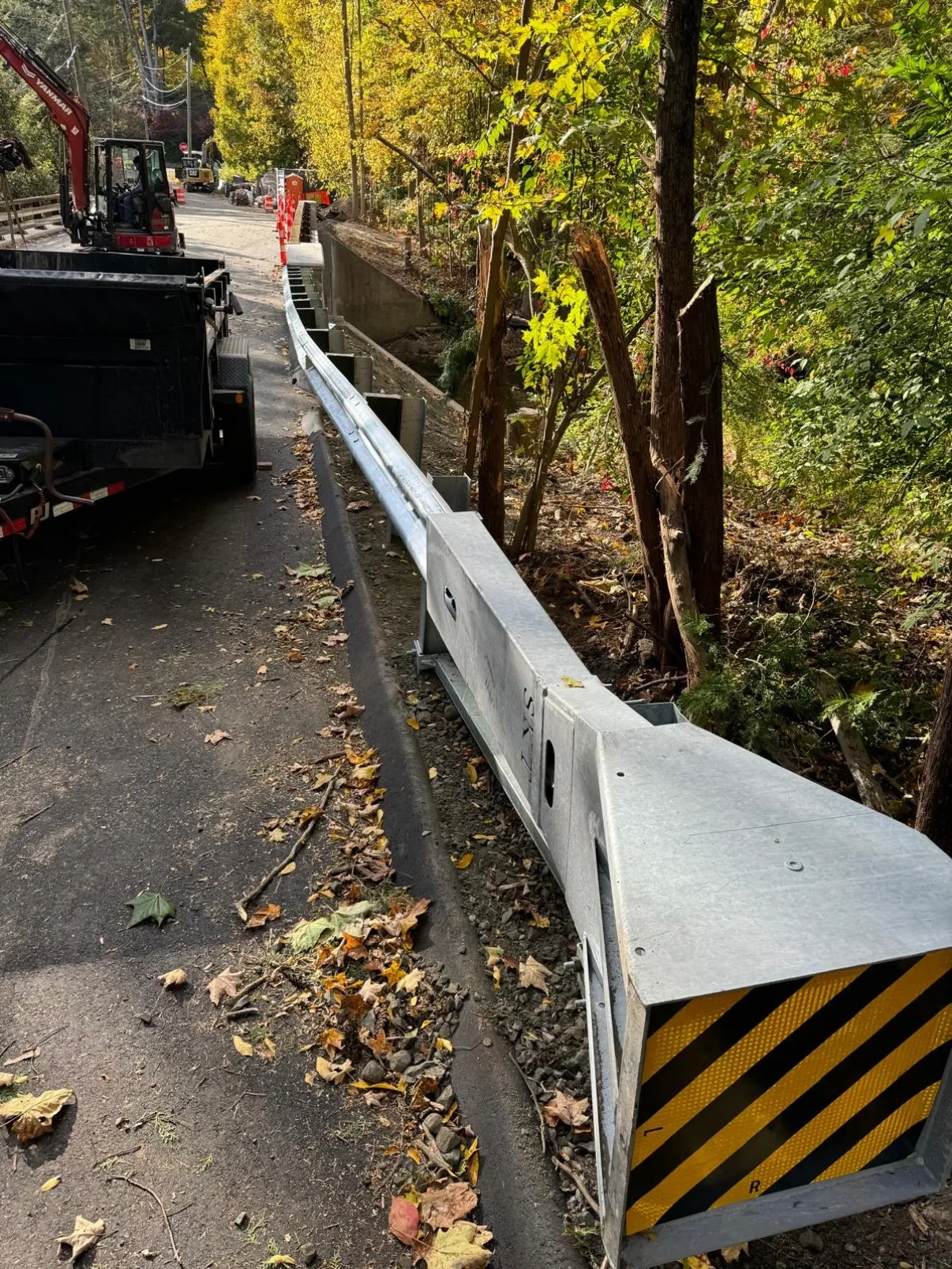 Newly installed galvanized W-beam guardrail with a yellow and black striped end terminal head along a wooded Connecticut roadside
