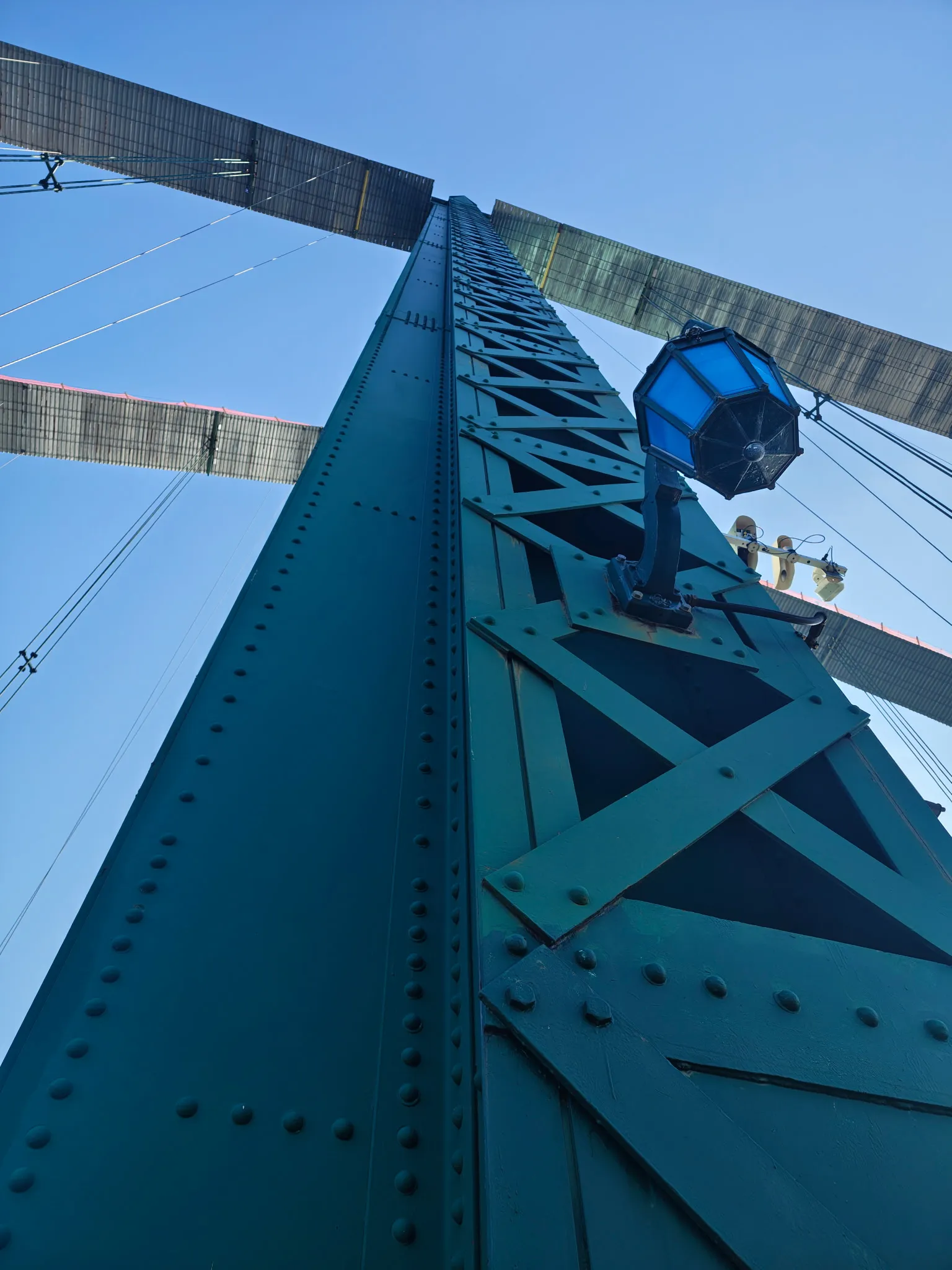 Freshly coated green steel tower of the Mount Hope Bridge seen from the deck looking straight up, showing riveted plate girders, cross-bracing, suspension cables and one of the bridge's octagonal blue lanterns