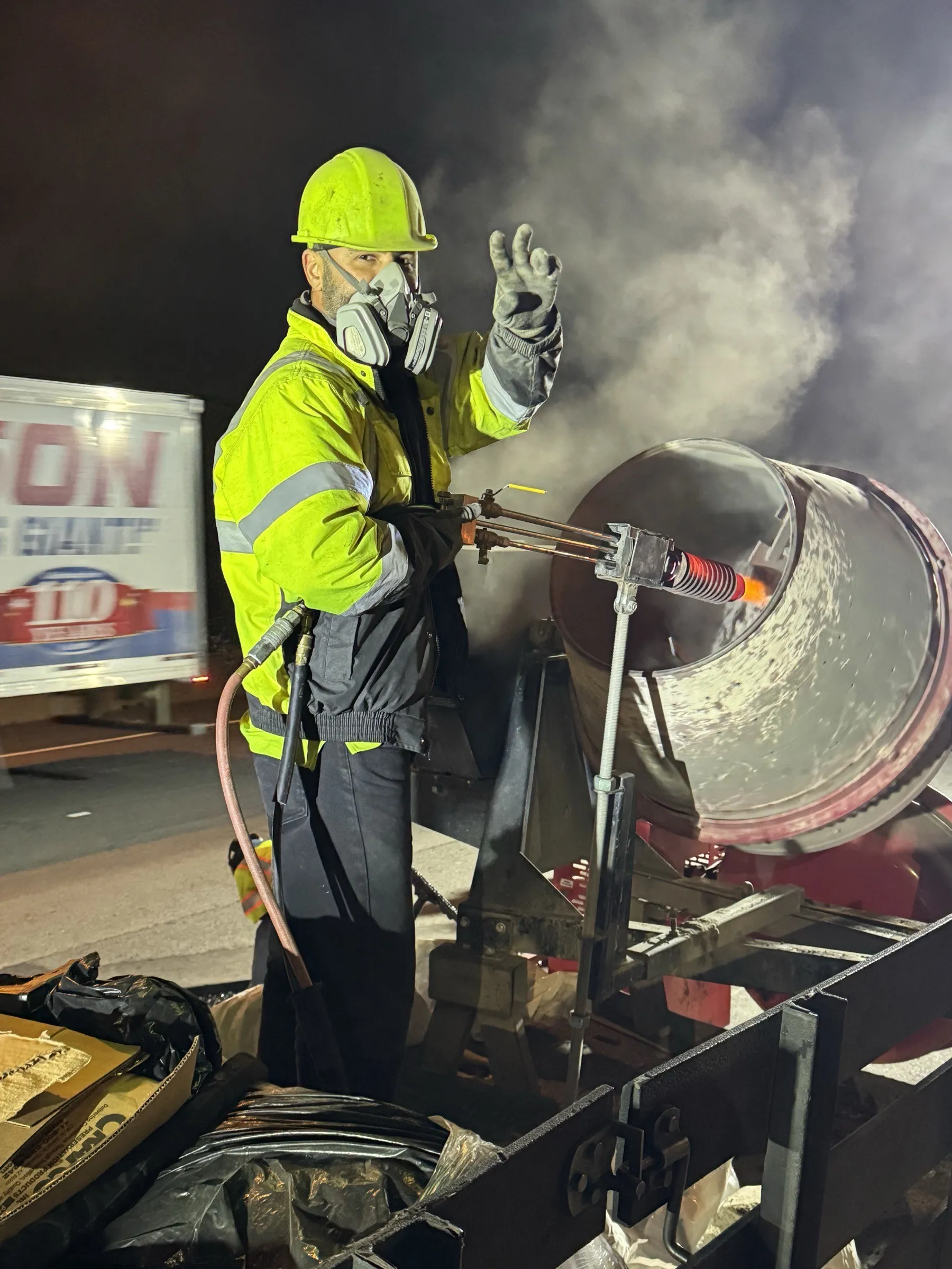 Cristiano & Son crewman in full PPE performing abrasive blasting on a bridge bearing at night, dust plume rising under floodlights