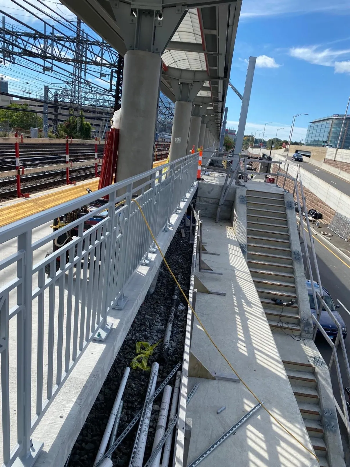 Commuter rail station platform under construction with new white steel picket railings, stair stringers, and canopy columns beside the tracks