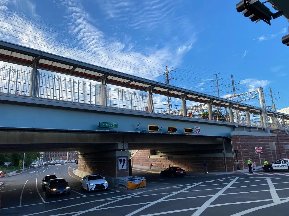 Railroad overpass carrying an elevated rail line above a city street, with tall protective fencing, platform canopy, and brick abutments